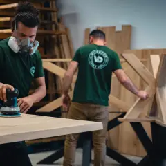 Un artisan porte un masque et utilise une ponceuse pour poncer du bois. Un autre artisan travaille sur un pied de table en bois.