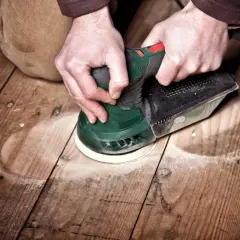 Mains tenant une ponceuse pour le ponçage du parquet. La poussière vole autour de l'outil pendant le travail.