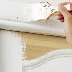 Main d'une personne peignant une porte en bois avec un pinceau. La peinture blanche recouvre partiellement le bois naturel. Quel rouleau pour peindre une porte en bois ?