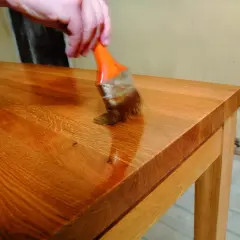 Application d'une huile dure sur un plateau en bois. Une main tient un pinceau orange pour appliquer le produit, révélant la beauté naturelle du bois. Recette huile dure.
