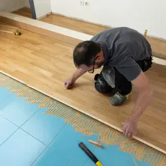 Un homme pose du parquet massif sur un sol bleu carrelé, appliquant de la colle. Il est en train de réaliser un projet de rénovation.