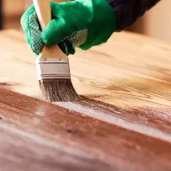 Application d'une couche de vernis sur un plateau en bois. La main gantée de vert applique le produit avec un pinceau, révélant la beauté naturelle du bois.