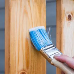 Application d'un produit pour protéger le bois extérieur. Une main tient un pinceau bleu et blanc pour appliquer le traitement sur des poteaux en bois clair.