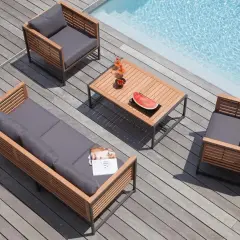 Salon de jardin en bois teck avec coussins gris, près d'une piscine. Table basse avec fruits et boissons.