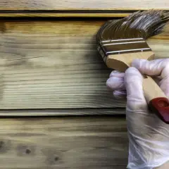 Maintenant, peut on mettre de la lasure sur de la peinture ? Un pinceau applique une lasure foncée sur un bardage en bois clair.