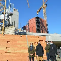 Trois hommes devant un mur en bois, un objet à faire au tour à bois, sur un chantier avec des grues.