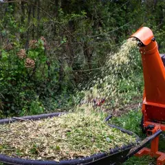 Une machine à faire des copeaux de bois orange projette des débris végétaux dans une brouette remplie de copeaux.