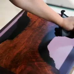 Main d'une personne polissant un plateau de table en bois et résine avec un chiffon bleu. Attention à la cire de carnauba danger.