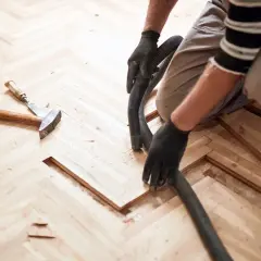 Un artisan utilise un aspirateur pour nettoyer la poussière de bois après le ponçage d'un parquet en chevrons. La pâte à bois blanchie au ponçage est prête pour la finition.