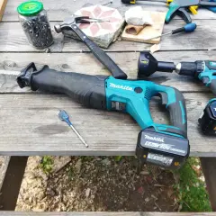 Outils de bricolage sur une table en bois : scie sabre et perceuse de marque makita, marteau, clous, mèche.