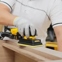 Main gantée utilise une ponceuse Mirka pour poncer une porte en bois, préparant la surface pour une finition parfaite.