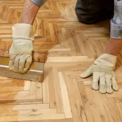 Un homme, ganté, applique de la cire sur un parquet en chevrons avec une raclette.