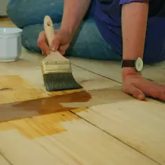 Application d'une couche protectrice sur un parquet huilé ou ciré. Une main tient un pinceau, l'autre est posée sur le bois.