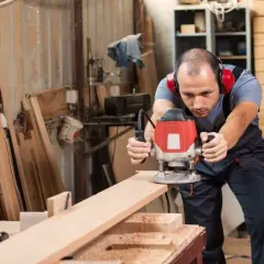 Artisan concentré sur ses réalisations à la défonceuse, créant des formes précises sur une planche de bois dans son atelier.