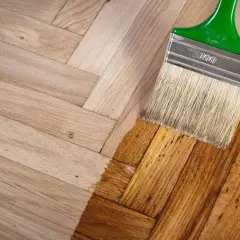 Un pinceau applique une couche de vernis sur un parquet en bois clair, transformant sa teinte. Le processus de nettoyer parquet est en cours.