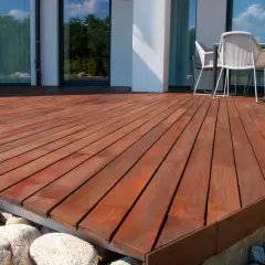 Belle terrasse bois sur plot béton, avec des galets et des gros rochers. Des chaises et une table sont visibles au fond.