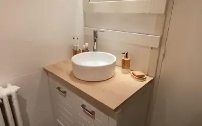 Un meuble de salle de bain relooké avec un plan de travail en bois clair, une vasque blanche et des accessoires modernes.