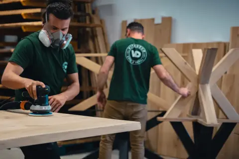 Un artisan porte un masque et utilise une ponceuse pour poncer du bois. Un autre artisan travaille sur un pied de table en bois.