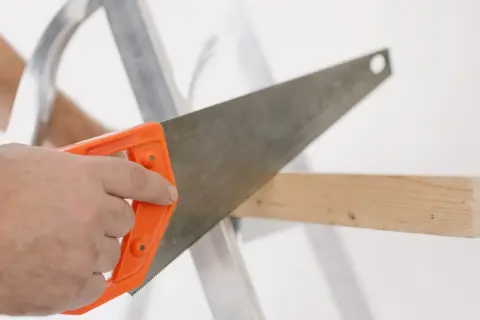 Main tenant une scie à bois à manche orange, coupant une planche. La différence scie à bois scie à métaux est dans la denture.