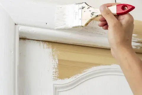 Main d'une personne peignant une porte en bois avec un pinceau. La peinture blanche recouvre partiellement le bois naturel. Quel rouleau pour peindre une porte en bois ?