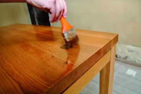 Application d'une huile dure sur un plateau en bois. Une main tient un pinceau orange pour appliquer le produit, révélant la beauté naturelle du bois. Recette huile dure.