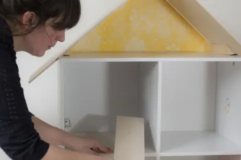 Une femme assemble une maison de poupée. Elle pose une pièce en bois dans une étagère blanche, sous un toit jaune à motifs.