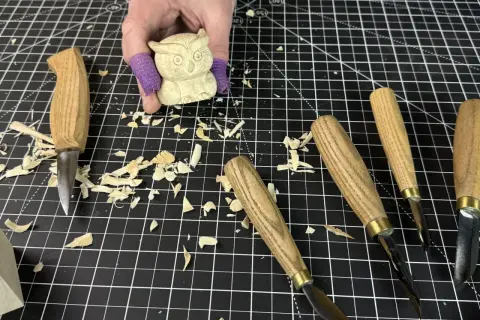 Une main tient une petite chouette en bois sculpté, entourée d'outils et de copeaux. Idéal pour une **débutant sculpture bois facile**.