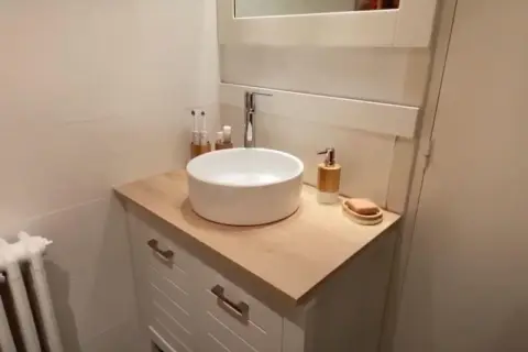 Un meuble de salle de bain relooké avec un plan de travail en bois clair, une vasque blanche et des accessoires modernes.
