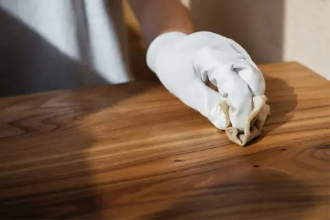 Main d'une personne portant un gant blanc appliquant de l'huile de lin dure naturelle sur un meuble en bois.