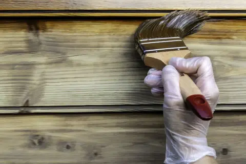 Maintenant, peut on mettre de la lasure sur de la peinture ? Un pinceau applique une lasure foncée sur un bardage en bois clair.