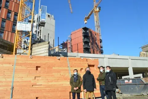 Trois hommes devant un mur en bois, un objet à faire au tour à bois, sur un chantier avec des grues.