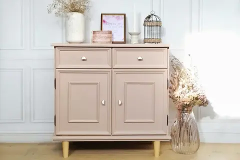 Un meuble en bois, fraîchement peint en rose poudré, est décoré de fleurs séchées et d'une cage à oiseaux.
