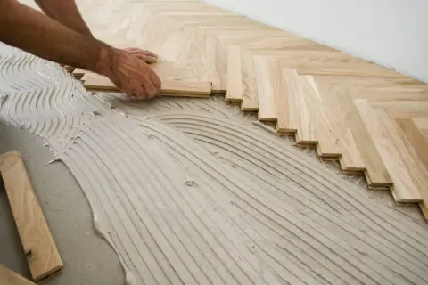 Pose d'un parquet en chevrons sur un lit de colle. Les lames de bois clair sont posées avec soin, formant un motif élégant.