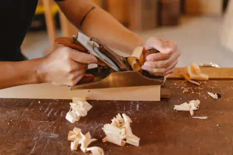 Main gauche tenant un rabot en laiton, créant des copeaux de bois. Une technique pour comment raboter du bois sans rabot.