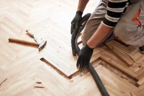 Un artisan utilise un aspirateur pour nettoyer la poussière de bois après le ponçage d'un parquet en chevrons. La pâte à bois blanchie au ponçage est prête pour la finition.