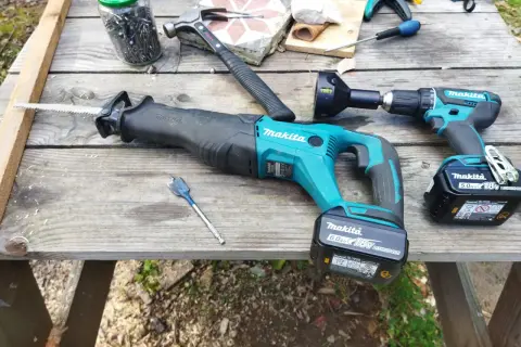 Outils de bricolage sur une table en bois : scie sabre et perceuse de marque makita, marteau, clous, mèche.