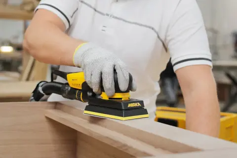 Main gantée utilise une ponceuse Mirka pour poncer une porte en bois, préparant la surface pour une finition parfaite.