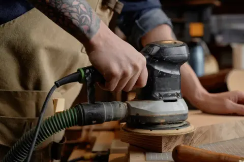 Main d'un artisan utilisant un outil pour poncer du bois. L'outil est relié à un aspirateur par un tuyau vert.