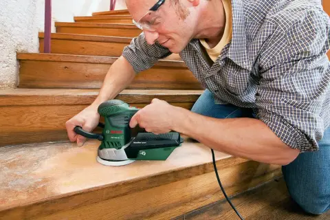 Un homme utilise une ponceuse pour rénover les marches d'un escalier en bois.
