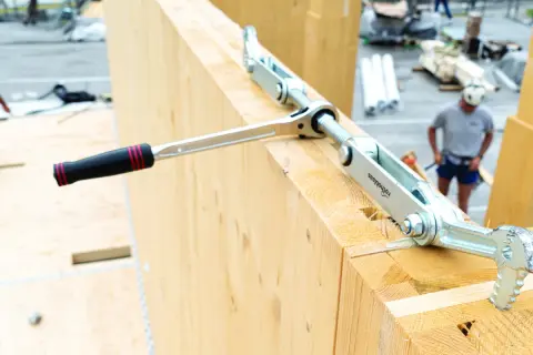 Un outil de serrage métallique, faisant partie d'une liste d'outils de charpentier, est utilisé sur de grosses poutres en bois.