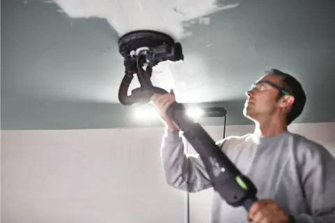 Un homme utilise une ponceuse de plafond Festool, éclairée par une lampe rasante Festool, pour lisser une surface.