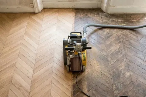 Ponçage d'un parquet ancien en chevrons, révélant sa beauté. Le prix du ponçage parquet dépend de l'état. Aspirateur industriel en action.