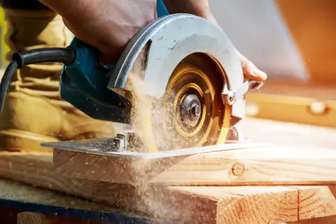 Main d'un artisan guidant une scie circulaire. Le sens de rotation de la lame de scie circulaire projette de la sciure.