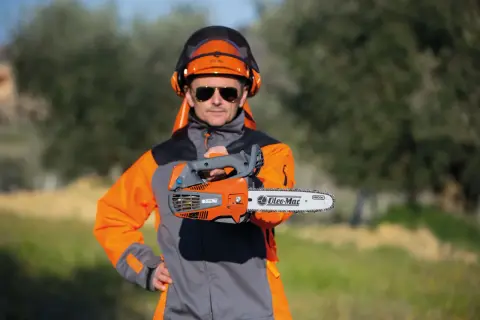 Un homme en tenue de protection, casque et lunettes, présente une tronçonneuse thermique. Il vous aide à choisir la meilleure.