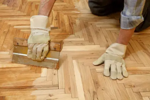 Un homme, ganté, applique de la cire sur un parquet en chevrons avec une raclette.