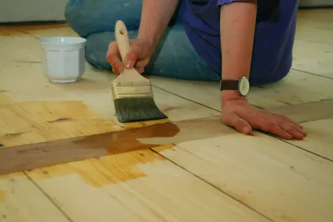 Application d'une couche protectrice sur un parquet huilé ou ciré. Une main tient un pinceau, l'autre est posée sur le bois.