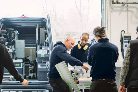 Des artisans discutent d'un outil Festool, l'origine de leur savoir-faire, devant un fourgon aménagé.