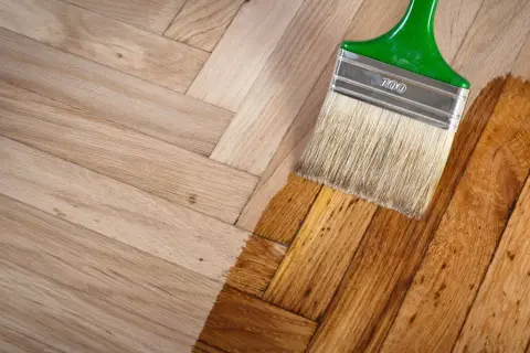 Un pinceau applique une couche de vernis sur un parquet en bois clair, transformant sa teinte. Le processus de nettoyer parquet est en cours.