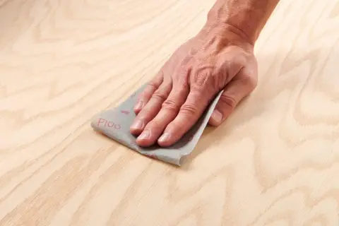 Main d'artisan effectuant un égrenage sur une surface en bois clair avec du papier de verre P100.