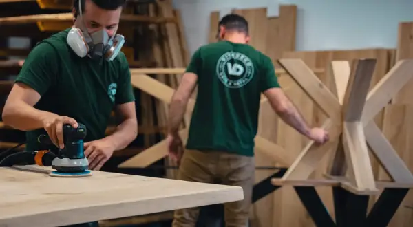 Un artisan porte un masque et utilise une ponceuse pour poncer du bois. Un autre artisan travaille sur un pied de table en bois.